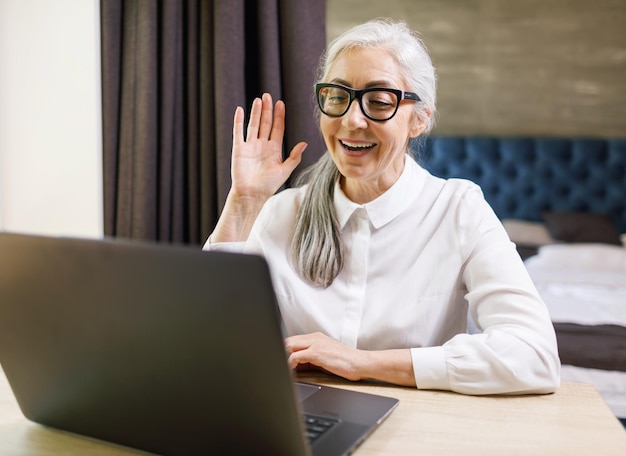 Senior donna dai capelli lunghi con gli occhiali che sventolano a mano mentre si utilizza il programma di videochat di famiglia sul laptop