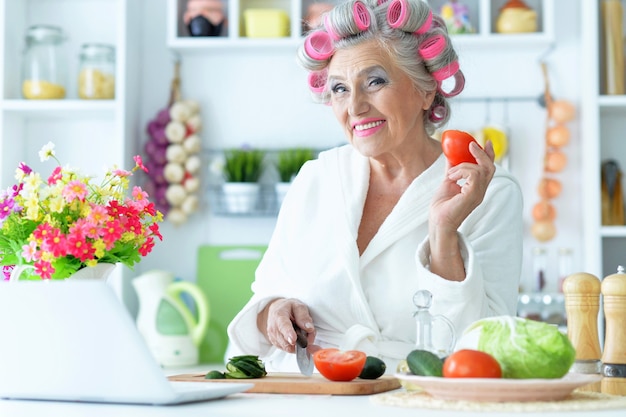 Senior donna in bigodini in cucina con laptop e verdure