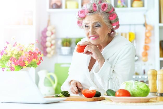 Senior donna in bigodini in cucina con laptop e verdure