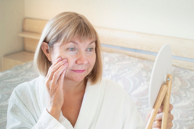 Senior donna in camera da letto applicando una lozione antietà. Guarda in uno specchio cosmetico. Concetto Anti età, sanità e cosmetologia, pensionato e maturo