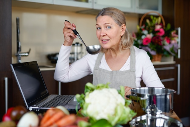 Senior donna in grembiule in cucina prepara il cibo e utilizza il computer portatile