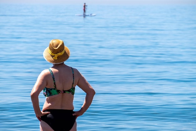 Senior donna in piedi da sola di fronte al mare mediterraneo su una spiaggia in una soleggiata giornata estiva