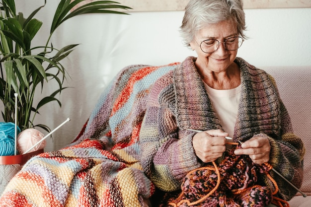 Senior donna matura seduta sul divano di casa mentre lavora a maglia e guardando il suo lavoro in corso Hobby pensionamento relax concetto per anziani sorridente donna nonna con gli occhiali