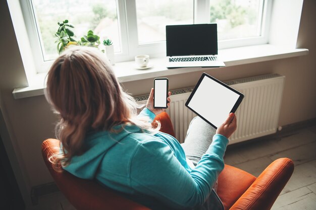 Senior donna seduta in poltrona con un tablet e un telefono mentre guarda il laptop