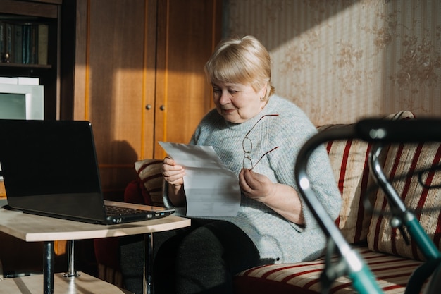 Senior donna utilizzando laptop e tenendo i documenti a casa. Femmina matura focalizzata che utilizza computer che lavora con prestiti bancari finanziari o documenti ipotecari online.