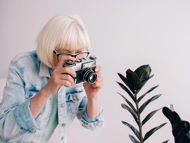 Senior elegante donna con capelli grigi con gli occhialigiacca di jeans per scattare una foto di fiori con la fotocamera a pellicola