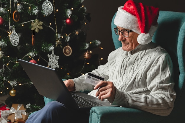 Senior in un cappello di Babbo Natale si siede su una poltrona utilizzando laptop tenendo la carta di credito sulla progettazione di un albero di Natale