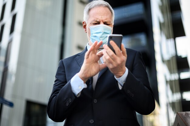 Senior manager che utilizza il suo smartphone all'aperto mentre indossa una maschera per proteggersi dalla pandemia di Coronavirus