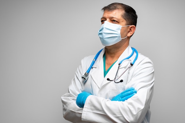 Senior maschio medico in maschera per il viso in piedi contro uno sfondo grigio