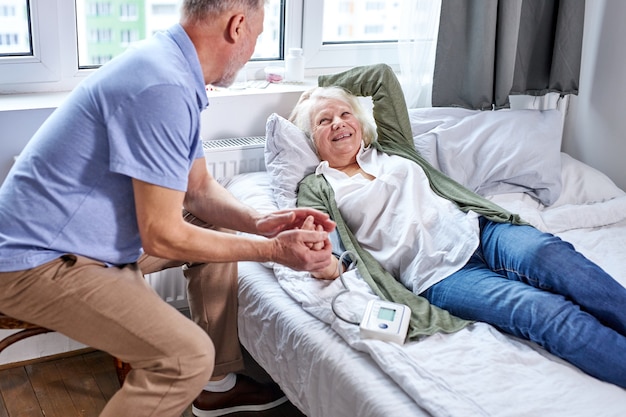 senior paziente femminile in ospedale con il marito preoccupato mano nella mano mentre si controlla la pressione sanguigna con il tonometro. l'uomo aiuta, supporto