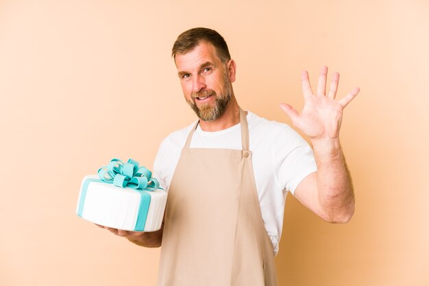 Senior tenendo una torta isolata sul muro beige sorridente allegro che mostra il numero cinque con le dita.