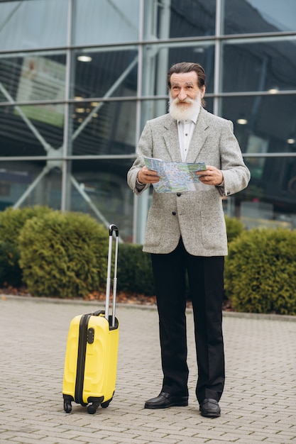 Senior uomo con la barba grigia con la valigia per il trasporto e la mappa della città sta camminando lungo l'edificio dell'aeroporto. Sta guardando da parte pensieroso. Copia spazio sul lato destro