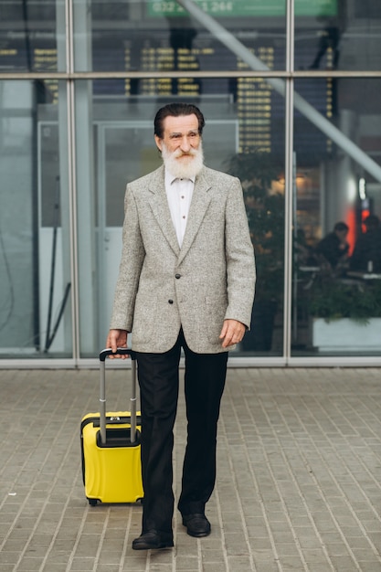 Senior uomo con la barba grigia con la valigia per il trasporto e la mappa della città sta camminando lungo l'edificio dell'aeroporto. Sta guardando da parte pensieroso. Copia spazio sul lato destro