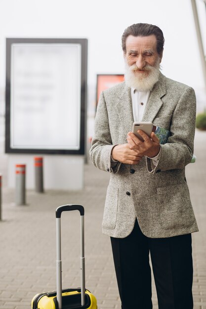 Senior uomo con la barba grigia con la valigia per il trasporto e la mappa della città sta camminando lungo l'edificio dell'aeroporto. Sta guardando da parte pensieroso. Copia spazio sul lato destro