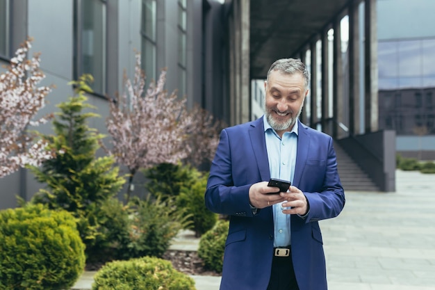 Senior uomo d'affari dai capelli grigi con esperienza fuori dall'ufficio sorridente e felice usando la lettura del capo del telefono