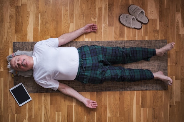 Senior uomo meditando su un pavimento di legno e sdraiato in posa Shavasana.