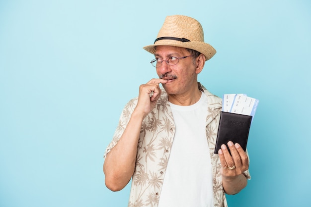 Senior viaggiatore indiano uomo di mezza età con passaporto isolato su sfondo blu rilassato pensando a qualcosa guardando uno spazio di copia.