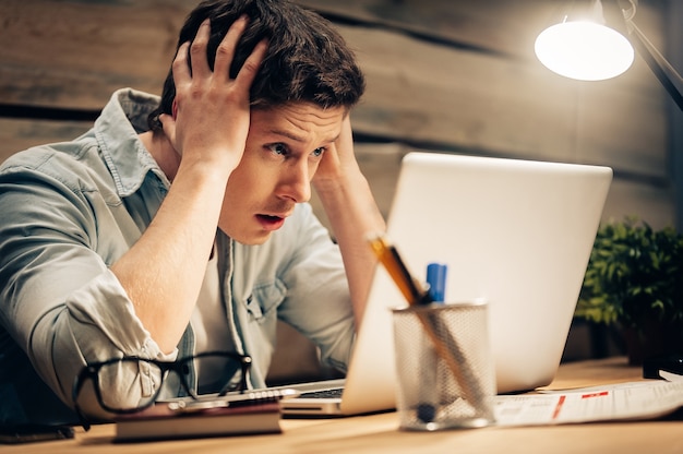 Sensazione di nausea e stanchezza. Giovane frustrato che tiene la testa tra le mani e guarda il laptop mentre lavora fino a tardi al suo posto di lavoro