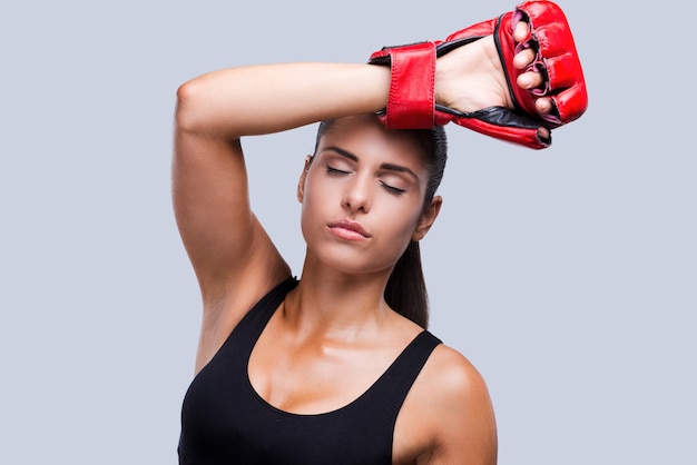 Sensazione di stanchezza dopo l'allenamento. Attraente giovane donna sportiva in guantoni da boxe che si tocca la fronte e tiene gli occhi chiusi mentre si trova in piedi su uno sfondo grigio