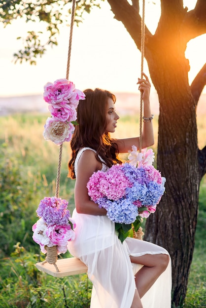 Sensuale bella ragazza con un sorriso perfetto in un abito bianco seduto su un'altalena in legno con un mazzo di teneri fiori colorati.