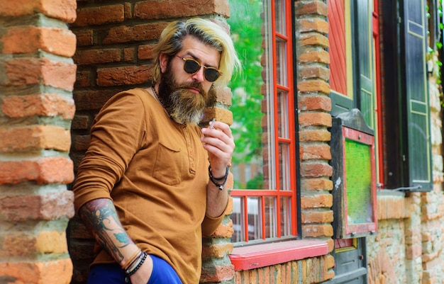 Sensuale uomo barbuto che fuma sigaretta elegante hipster in occhiali da sole fuma tabacco da sigaretta