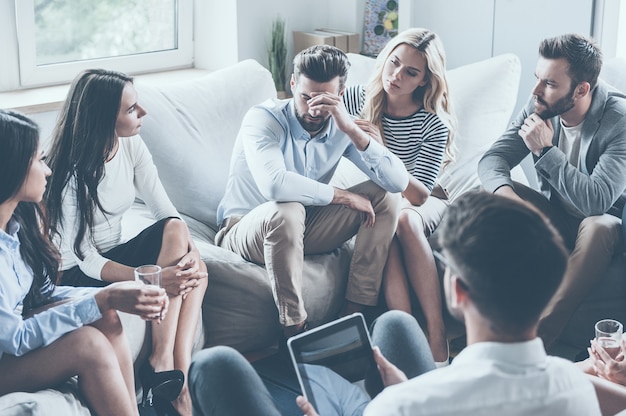 Sentirsi depresso. Gruppo di giovani seduti in cerchio mentre un uomo tiene la testa in mano e sembra sconvolto mentre una giovane donna lo conforta