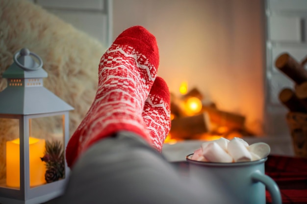 Serata d'inverno accogliente a casa, piedi in caldi calzini rossi di lana accanto al caminetto, una donna è rilassante e con una tazza di bevanda calda. Atmosfera natalizia.