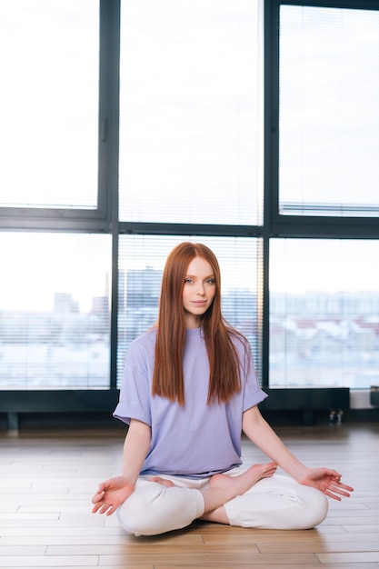 Serena attraente giovane donna meditando seduta sul pavimento nella posa del loto sullo sfondo della finestra nella stanza dell'ufficio luminoso.