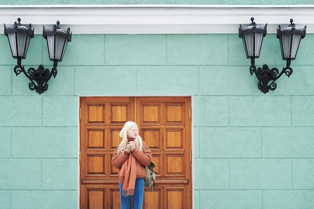 Serena ragazza albina in blue jeans e giacca marrone in piedi accanto alla porta d'ingresso