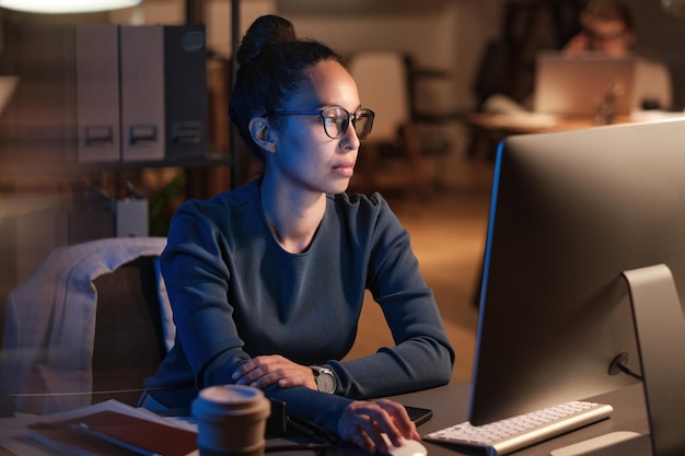 Seria giovane donna d'affari con gli occhiali seduta al tavolo e utilizzando il computer mentre prepara la presentazione di notte