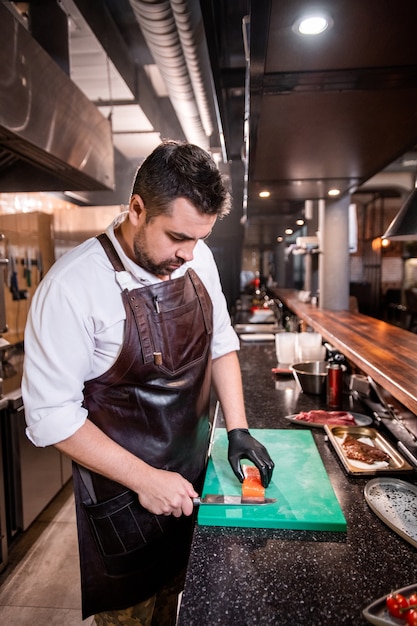 Serio chef impegnato in grembiule in pelle in piedi al bancone e prepara bistecca di pesce per cucinare il piatto nel ristorante