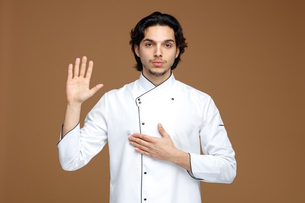 serio giovane chef maschio che indossa l'uniforme guardando la fotocamera che mostra il gesto di promessa isolato su sfondo marrone