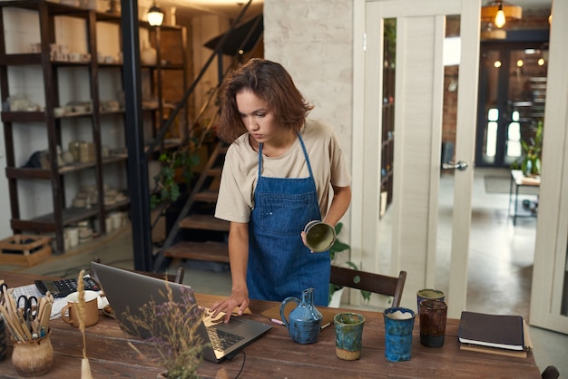 Serio giovane proprietario caucasico di un negozio di ceramiche in grembiule di jeans che tiene in mano una tazza artigianale e usa il laptop mentre...