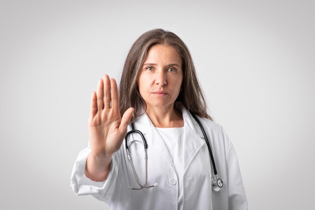 Serio medico donna senior europea in camice bianco con stetoscopio che mostra il segnale di stop alla fotocamera grigia