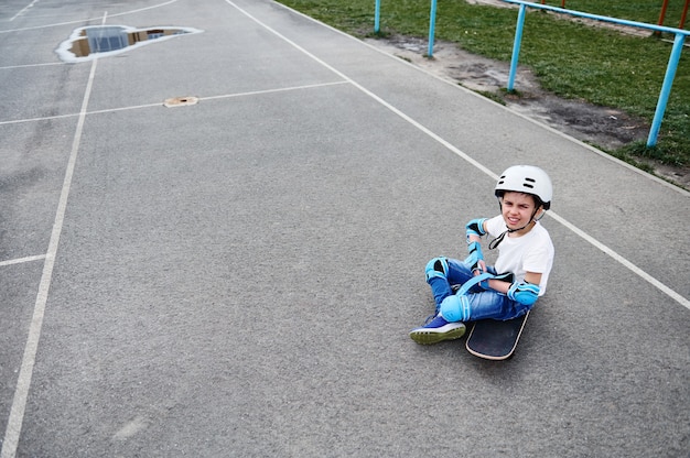 Serious oy nel casco di sicurezza si siede sullo skateboard e si concentra sull'indossare indumenti protettivi per lo skateboarder