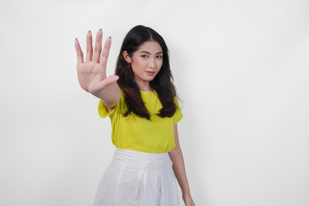 Serious young Asian woman in a yellow shirt and white skirt is holding up her hand to make a stop sign
