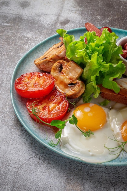 Serve un piatto dal menu del ristorante. Deliziosa e sana colazione con uova fritte, fagioli, salsicce, verdure grigliate, pomodori e funghi con insalata su un piatto contro un tavolo di pietra grigia