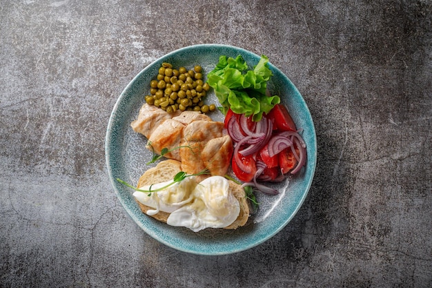 Serve un piatto dal menu del ristorante. Petto di pollo con verdure e verdure, pomodori, cipolle, omelette e piselli su un piatto sullo sfondo di un tavolo in pietra grigia