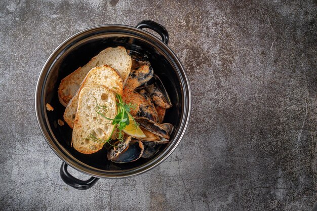 Serve un piatto dal menu di un ristorante. Cozze fritte con crostini e limone in una padella scura contro un tavolo di pietra grigia