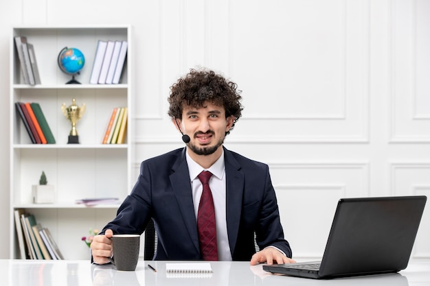 Servizio clienti bel ragazzo in tuta da ufficio con laptop e auricolare sorridente con il caffè