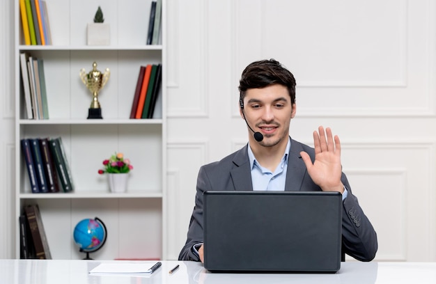 Servizio clienti bell'uomo in abito grigio con computer e auricolare che dice ciao al cliente
