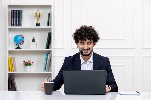 Servizio clienti bell'uomo riccio in tuta da ufficio con computer e auricolare con tazza di caffè