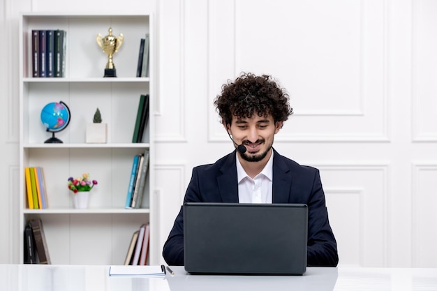 Servizio clienti carino ragazzo bruna in tuta da ufficio con laptop e auricolare funzionante e sorridente