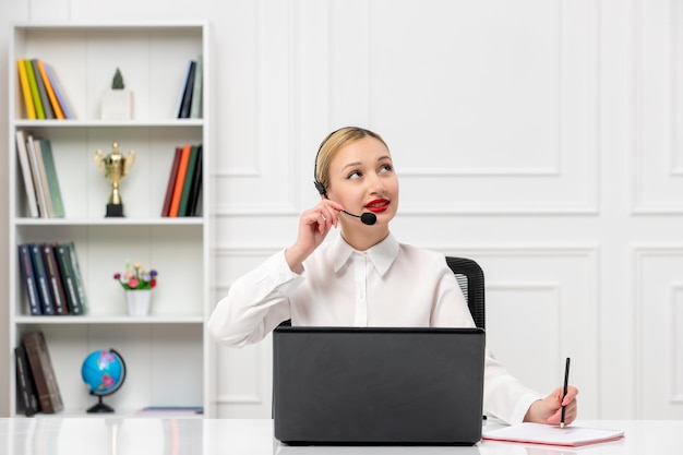 Servizio clienti ragazza bionda carina in camicia da ufficio con auricolare e computer alzando lo sguardo e pensando