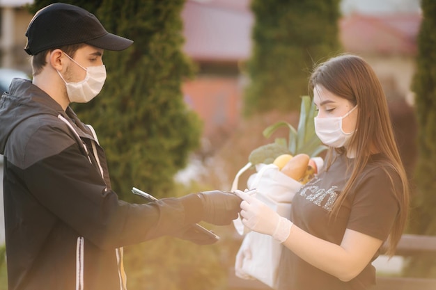 Servizio di consegna di cibo intelligente Uomo in maschera medica e guanti che consegnano cibo fresco a una giovane donna cliente