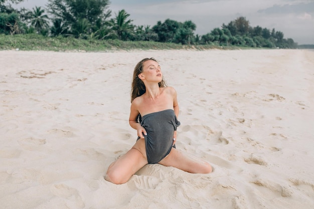 Servizio fotografico sulla spiaggia di sabbia. Una giovane bella ragazza posa seducente seduta sulla sabbia in un body grigio leggermente rimosso con gli occhi chiusi. Sfondo tropicale con alberi e cielo.