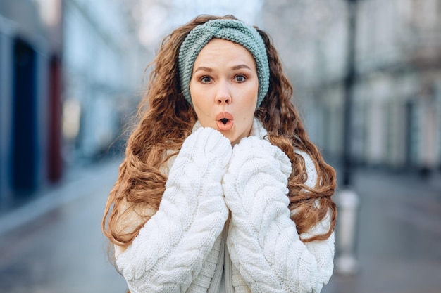 Sessione fotografica emozionale per le strade della città. Giovane ragazza in abiti caldi per il clima fresco pone in piedi mostrando sorpresa con la bocca rotonda aperta e gli occhi sorpresi. Il concetto di stato di shock.