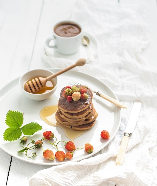 Set colazione. Frittelle di grano saraceno con fragole fresche di giardino, miele e tazza di caffè sul tavolo di legno bianco