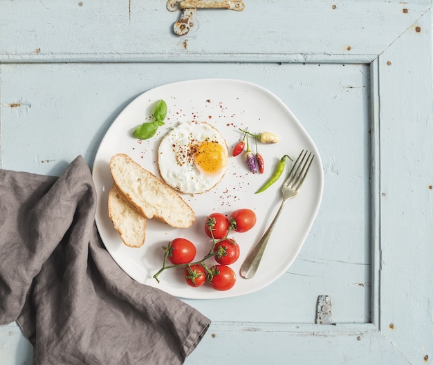 Set colazione. Uovo fritto, fette di pane, pomodori ciliegia, peperoncini ed erbe sul piatto ceramico bianco sopra la tavola di legno blu-chiaro, vista superiore.