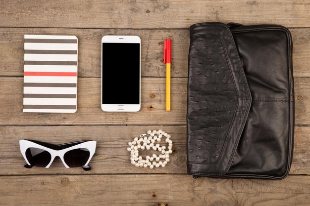 Set da donna con borsa per smartphone, occhiali da sole, blocco note, penna e perla su scrivania in legno marrone